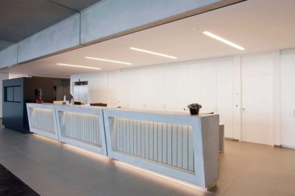 Empfangstheke mit Lichtband vor weißer Wandverkleidung mit Einbauschränken – Willy Naessens Group, Belgien