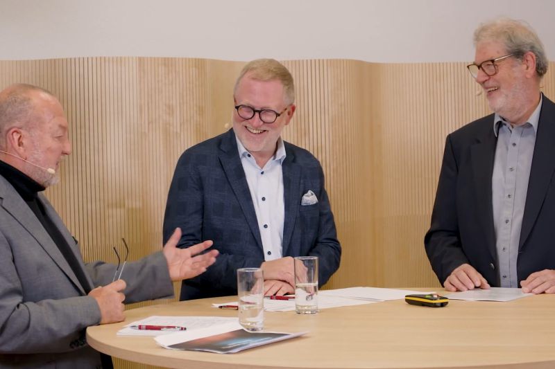 Drei Männer diskutieren lebhaft über Raumakustik vor einer Wandverkleidung aus Lochplatten in einem modernen Raum.