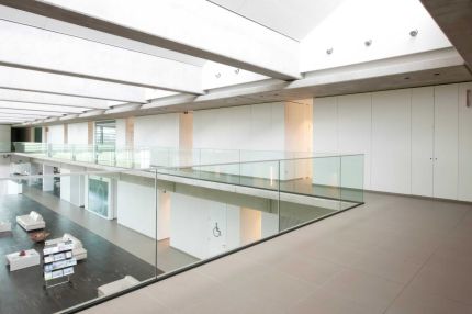 Galerie mit Glasbrüstung über dem Atrium, weiße Wandverkleidung und Lounge darunter – Willy Naessens Group, Belgien