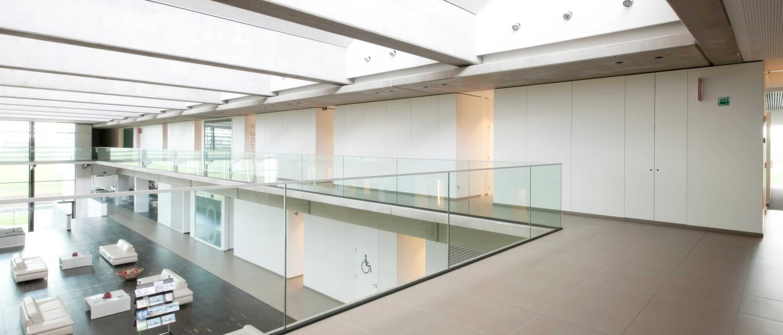 Galerie mit Glasbrüstung über dem Atrium, weiße Wandverkleidung und Lounge darunter – Willy Naessens Group, Belgien