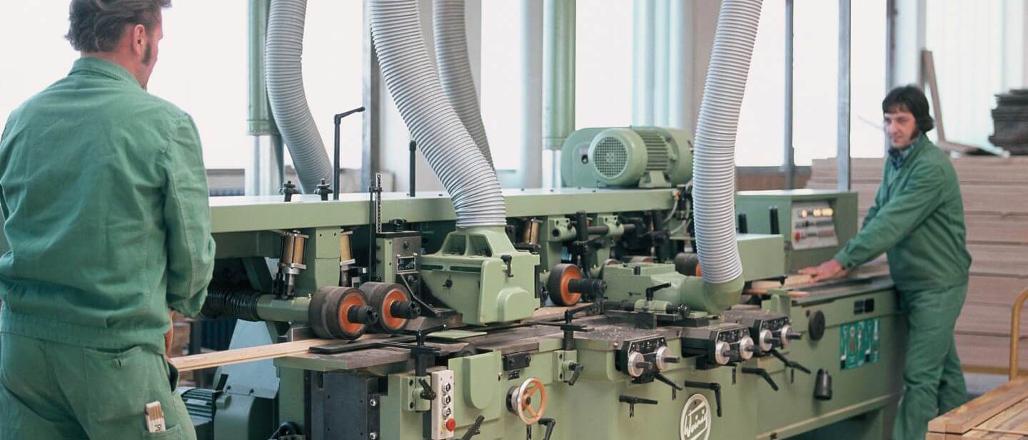 Zwei Arbeiter in grünen Overalls bedienen eine Maschine zur Bearbeitung von Holz für akustische Wandverkleidungen.