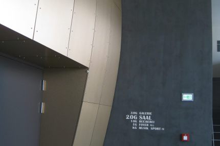 Foyer mit bronzefarbener Wandverkleidung neben dunkler Wand mit Geschossübersicht – Bürgerhaus Lechner Bräu