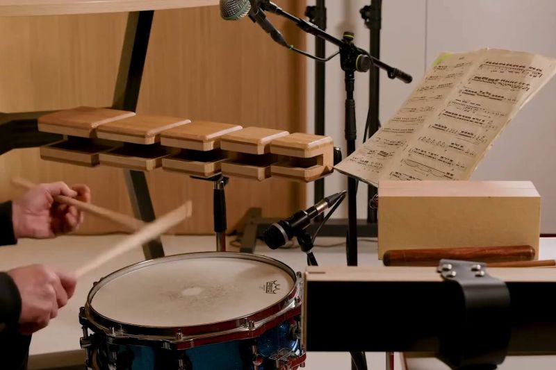 Ein Musiker spielt auf einer Trommel und einem Xylophon, während Notenblätter und Mikrofone im Raum für optimale Raumakustik sorgen.