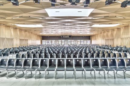 Saal mit Stuhlreihen, facettierter Holzdecke und Bühnenscheinwerfern – Gymnasium Oberursel