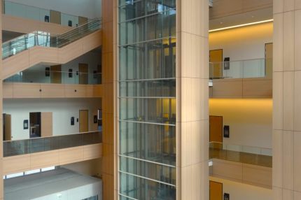 Gläserner Aufzugsschacht zwischen holzverkleideten Pfeilern im Lichthof – Justizzentrum Bochum