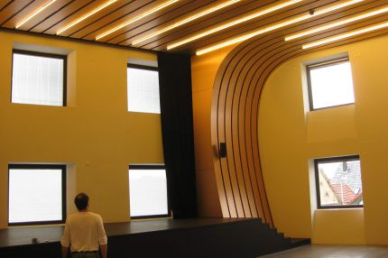 Saal mit Bühne, Holzlamellendecke mit Lichtbändern und gelben Wänden – Bürgerhaus Lechner Bräu