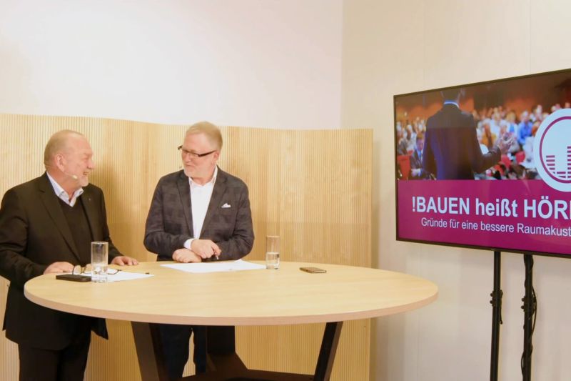 Zwei Redner diskutieren am Tisch über Raumakustik, während auf dem Bildschirm Lösungen für akustische Herausforderungen angezeigt werden.