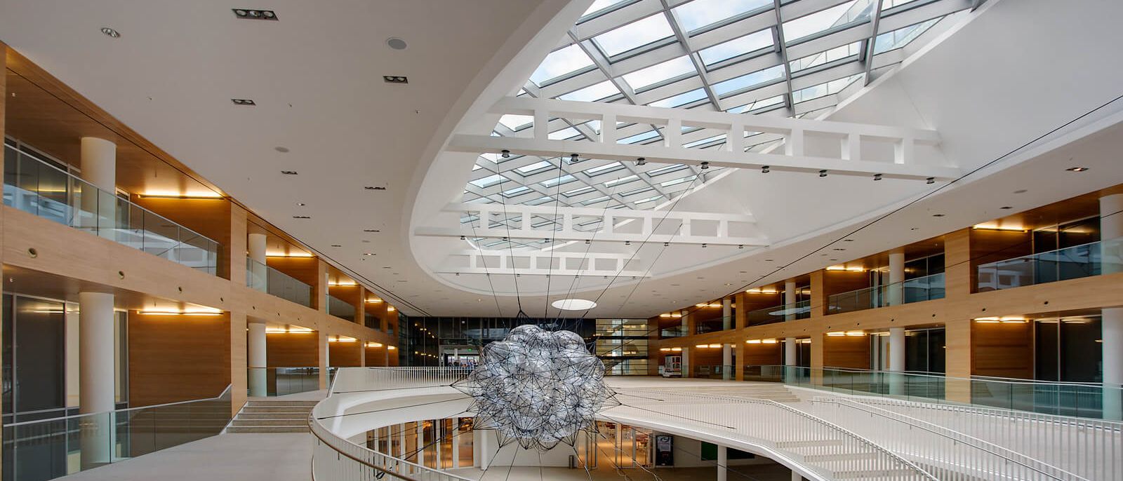 Halle unter ovalem Glasdach mit hängender Skulptur und holzverkleideten Galerien – Europäisches Patentamt