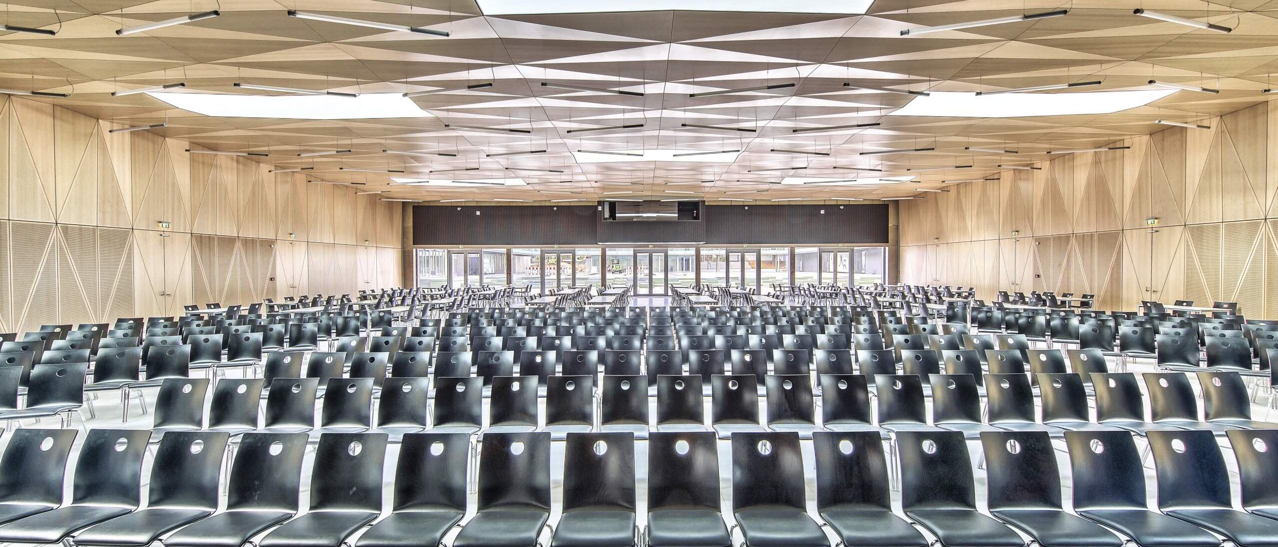 Saal mit Stuhlreihen, facettierter Holzdecke und Bühnenscheinwerfern – Gymnasium Oberursel