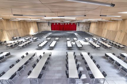 Mehrzwecksaal von oben mit Tischreihen, Holz-Wandverkleidung und rotem Bühnenvorhang – Gymnasium Oberursel