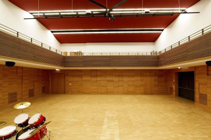 Saal mit Parkett, Holz-Wandverkleidung, Lamellengalerie und roter Decke – Musikschule Essenbach