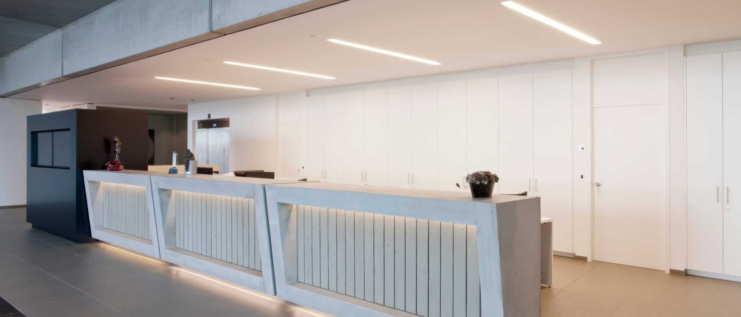 Empfangstheke mit Lichtband vor weißer Wandverkleidung mit Einbauschränken – Willy Naessens Group, Belgien
