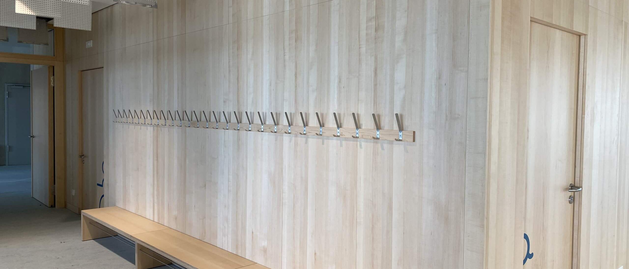 Flur mit Holz-Wandverkleidung, Garderobenhaken, Sitzbank und Akustikdecke – Bildungskampus Köln