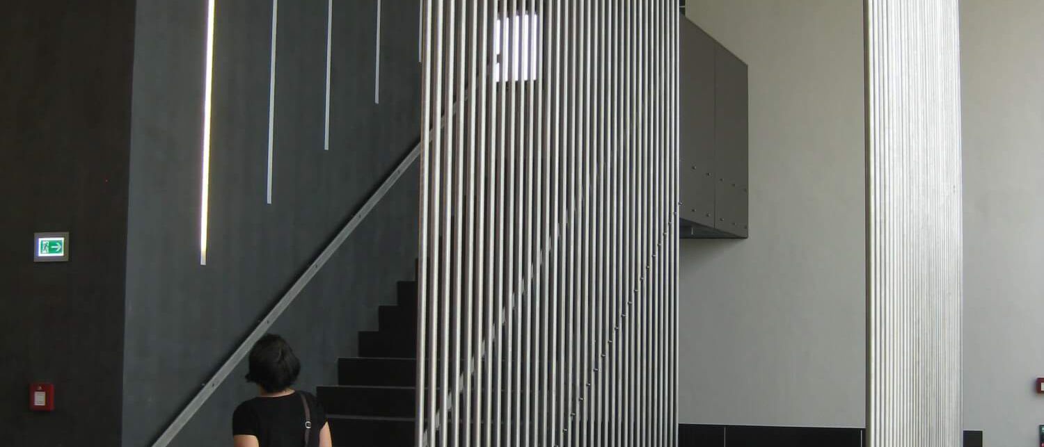 Treppenhaus mit dunkler Wand, Lichtschlitzen und Edelstahl-Stabgitter – Bürgerhaus Lechner Bräu
