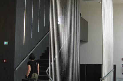Treppenhaus mit dunkler Wand, Lichtschlitzen und Edelstahl-Stabgitter – Bürgerhaus Lechner Bräu