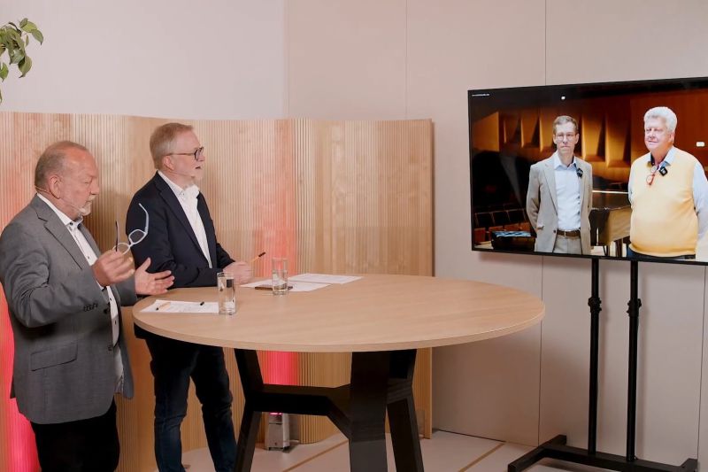 Zwei Männer diskutieren am Tisch über Raumakustik, während eine Präsentation mit akustischen Lösungen auf dem Bildschirm gezeigt wird.