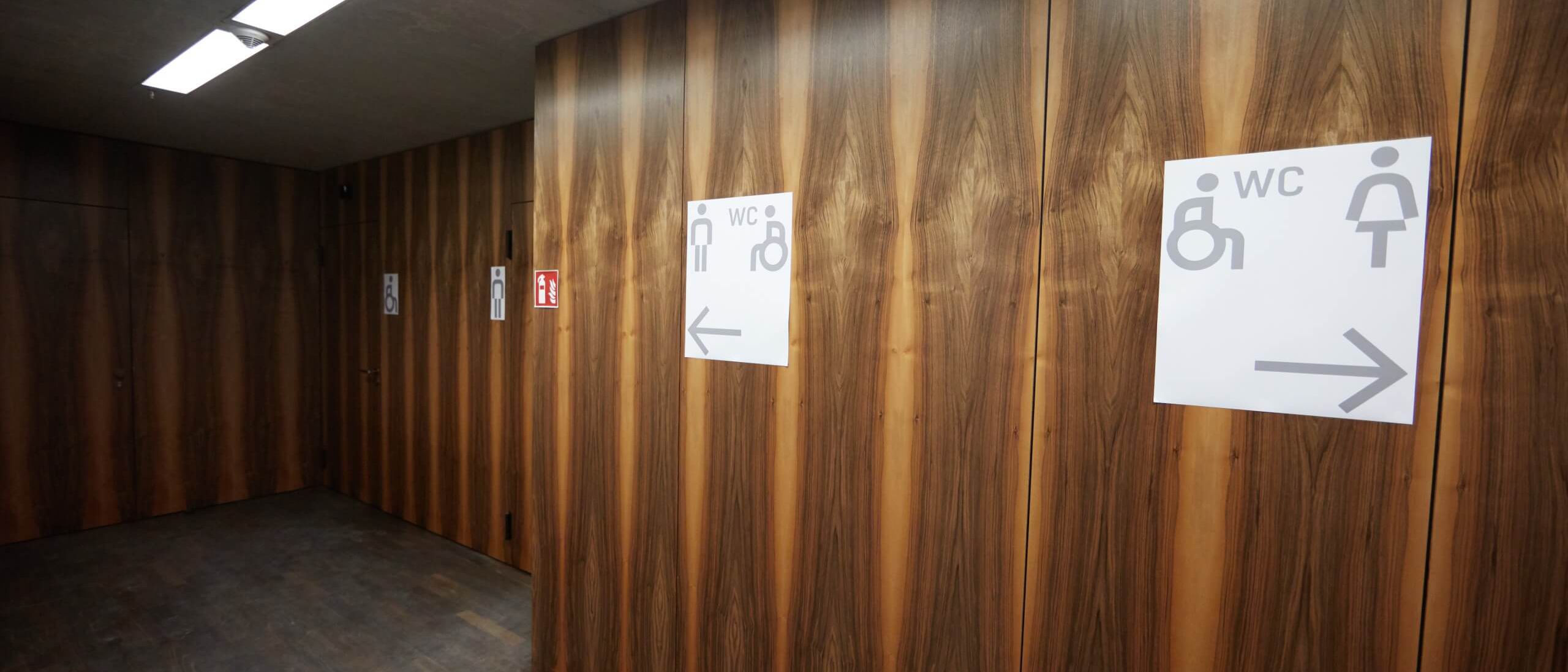 Vorraum der Toiletten mit Wandverkleidung in Nussbaum-Furnier und Beschilderung – James-Simon-Galerie, Berlin