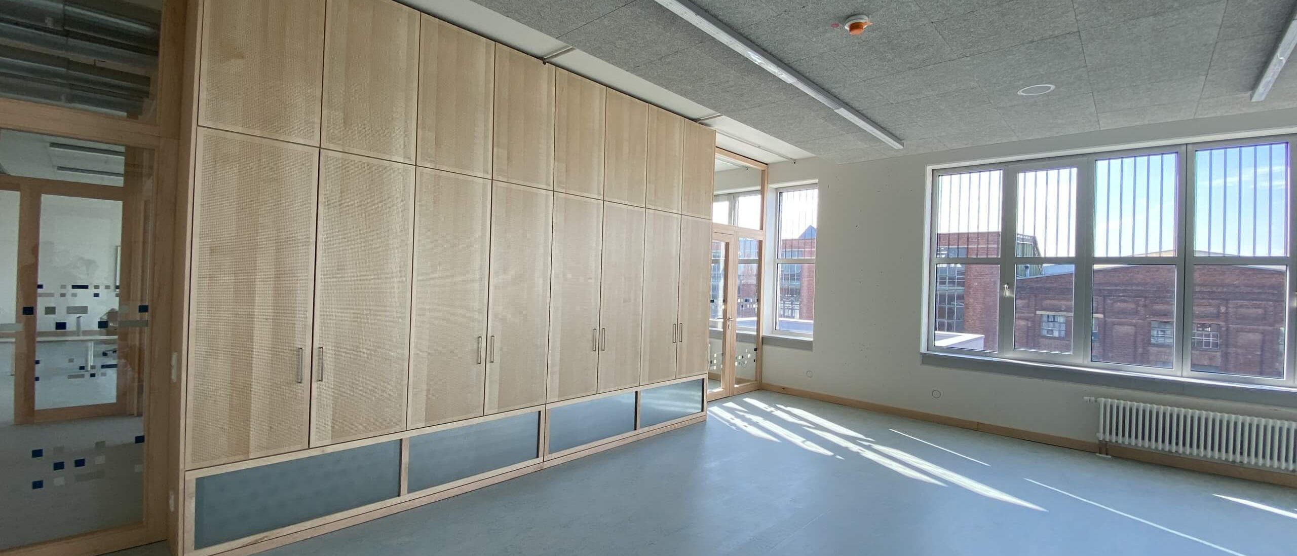 Klassenraum mit Einbauschrank aus Holz, Akustikdecke und großen Fenstern – Bildungskampus Köln