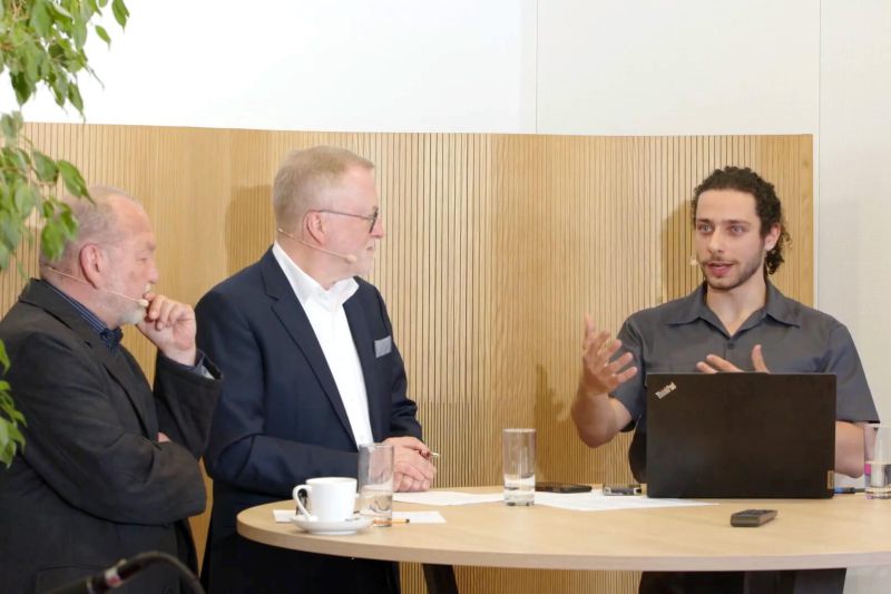 Ein Presenter diskutiert mit zwei Kollegen über innovative Akustiklösungen, während stilvolle Lochplatten im Hintergrund hervorgehoben werden.