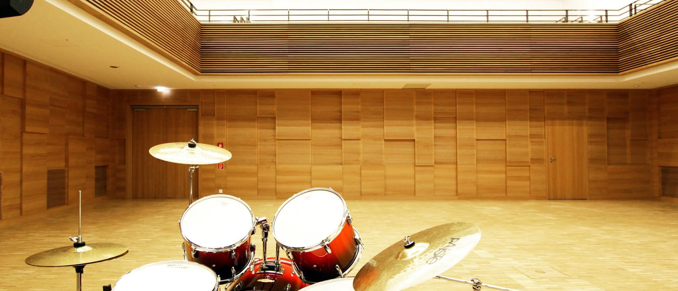 Saal mit Schlagzeug, Holz-Wandverkleidung und Galerie mit Lamellenbrüstung – Musikschule Essenbach