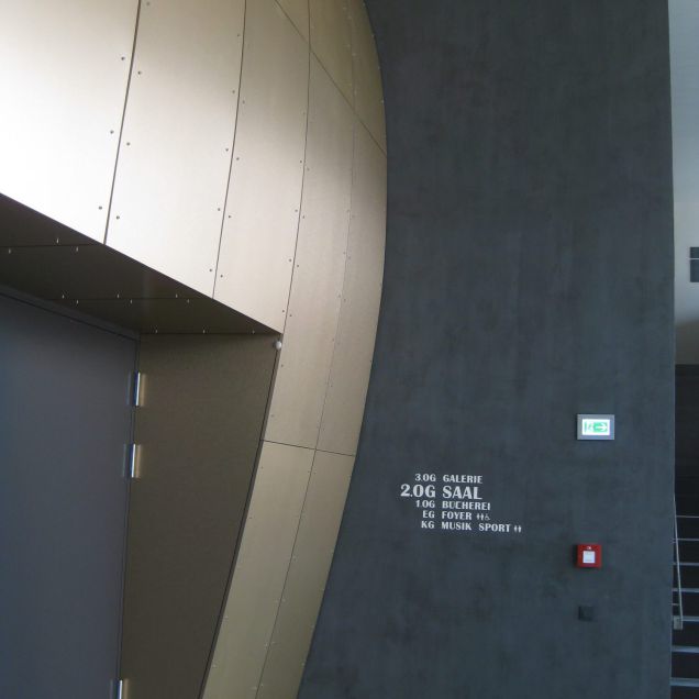 Foyer mit bronzefarbener Wandverkleidung neben dunkler Wand mit Geschossübersicht – Bürgerhaus Lechner Bräu
