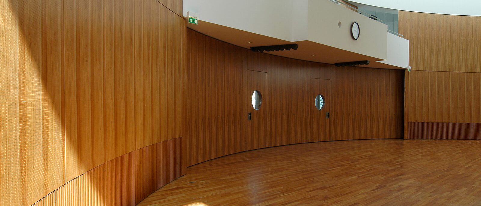 Leerer Saal mit gebogener Holz-Wandverkleidung und Bullaugentüren unter der Empore – IHK Potsdam