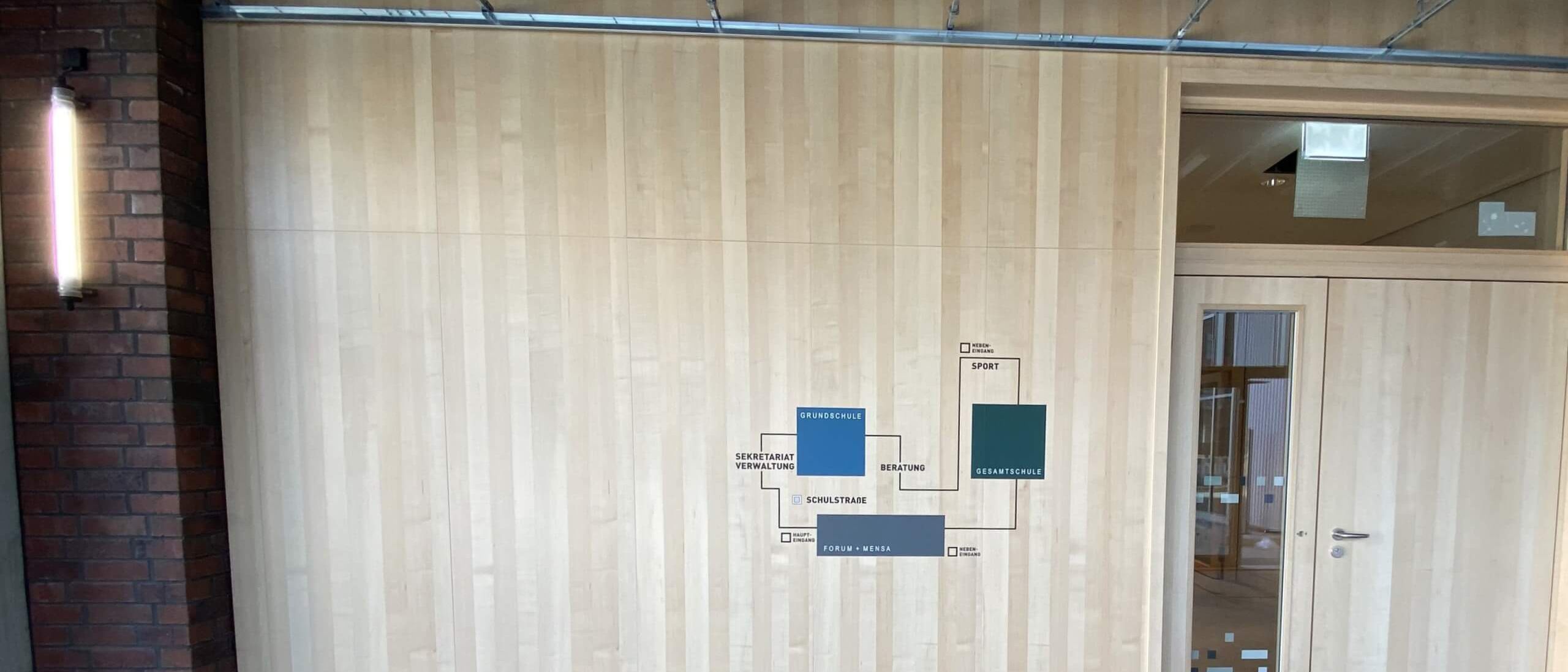 Holz-Wandverkleidung mit Orientierungsplan neben Klinkerpfeiler und Doppeltür – Bildungskampus Köln