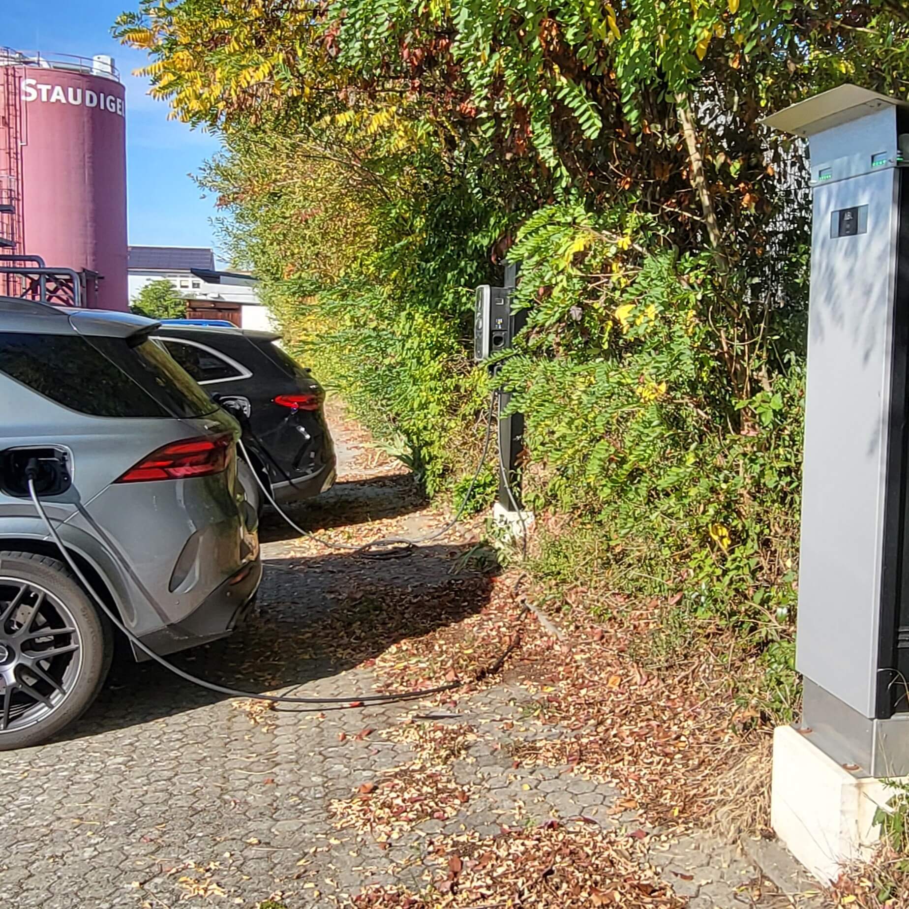 Zwei Elektroautos laden an Ladesäulen in der Nähe von Staudigel unter grünen Bäumen.